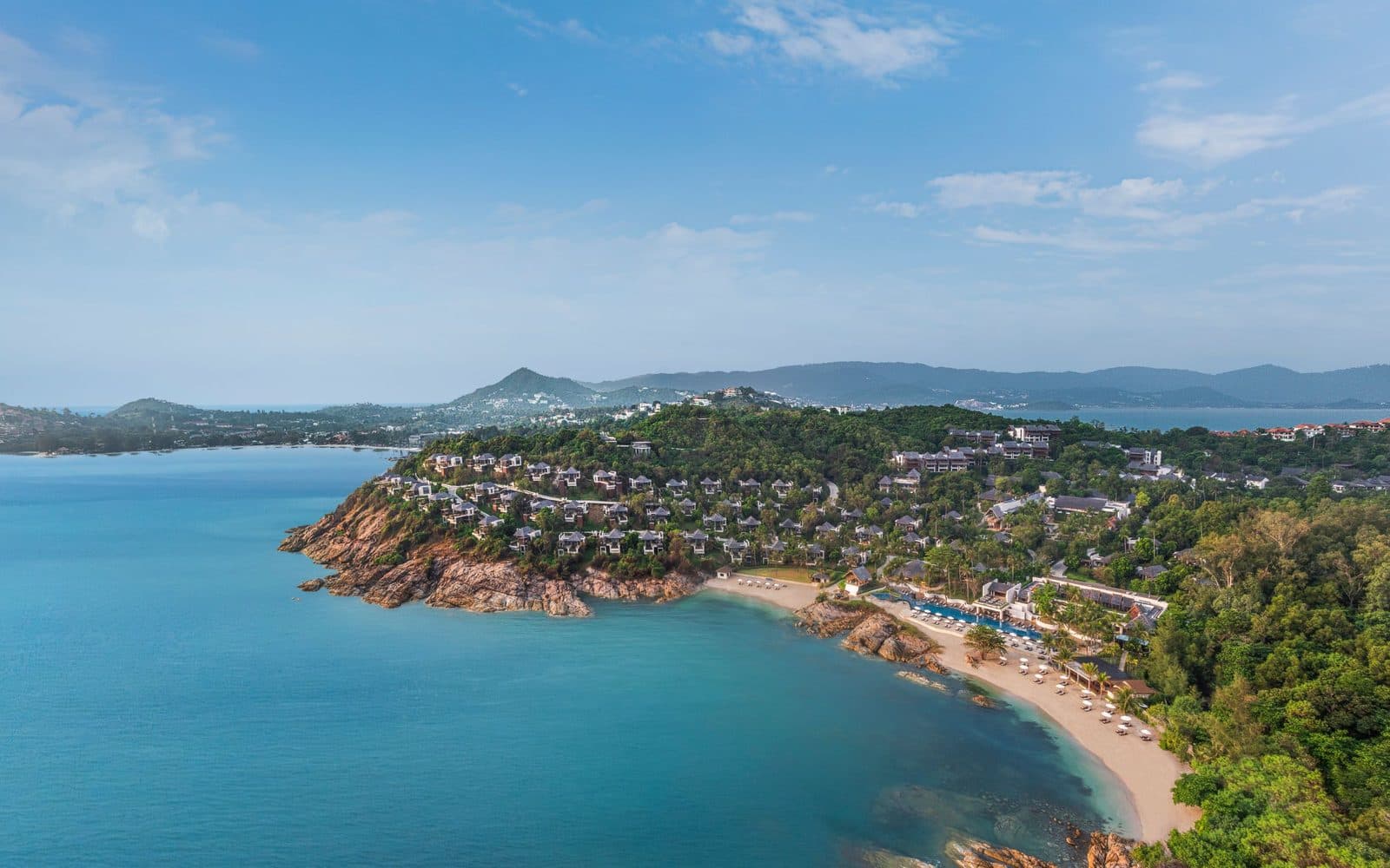 Вид сверху на The Ritz-Carlton, Koh Samui: виллы на скалистом мысу, пляж с бассейном у бухты и горы Самуи вдали