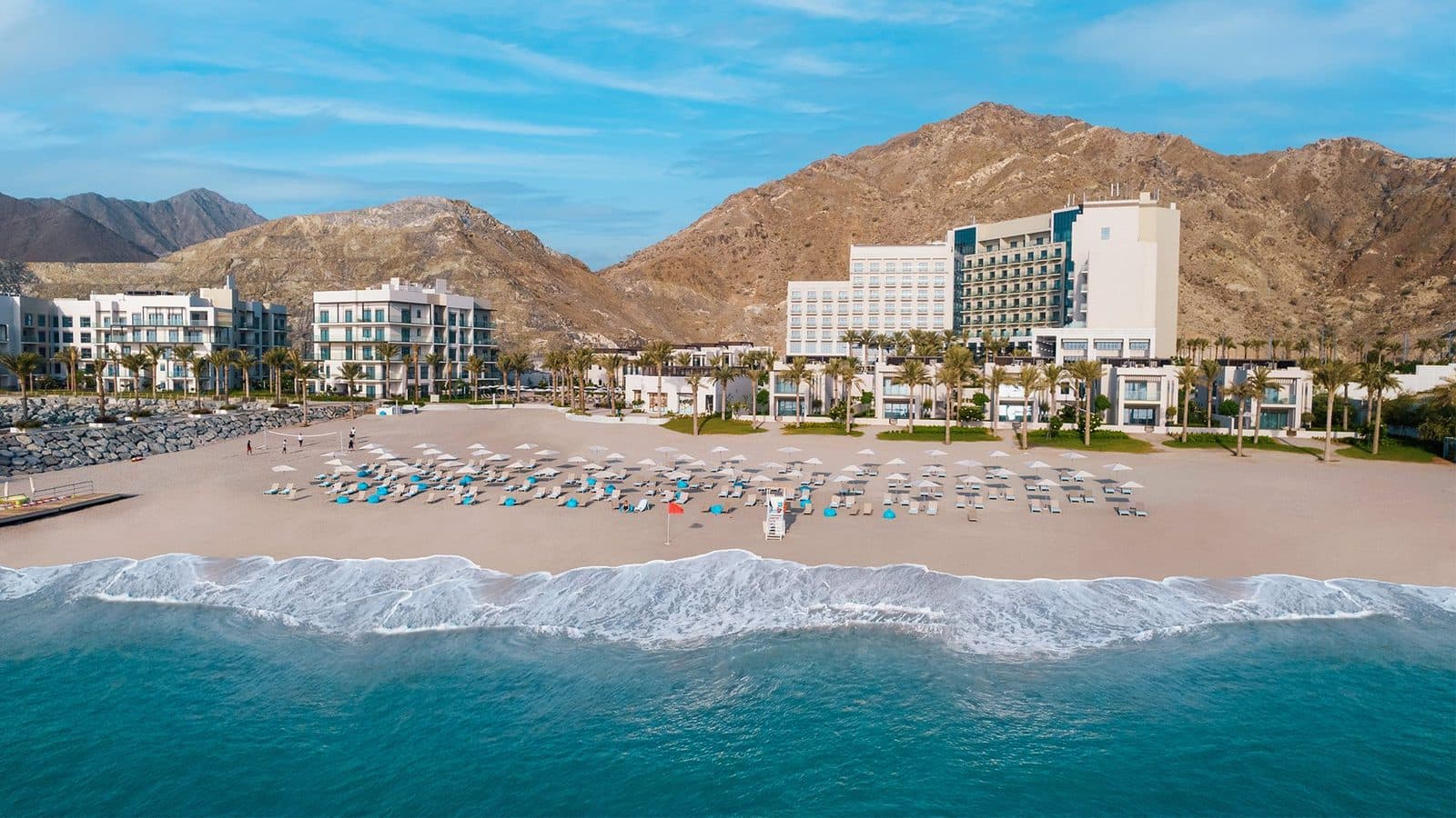 Вид с моря на Address Beach Resort Fujairah: широкий пляж с зонтиками и шезлонгами, белые корпуса отеля среди пальм и горы Хаджар позади