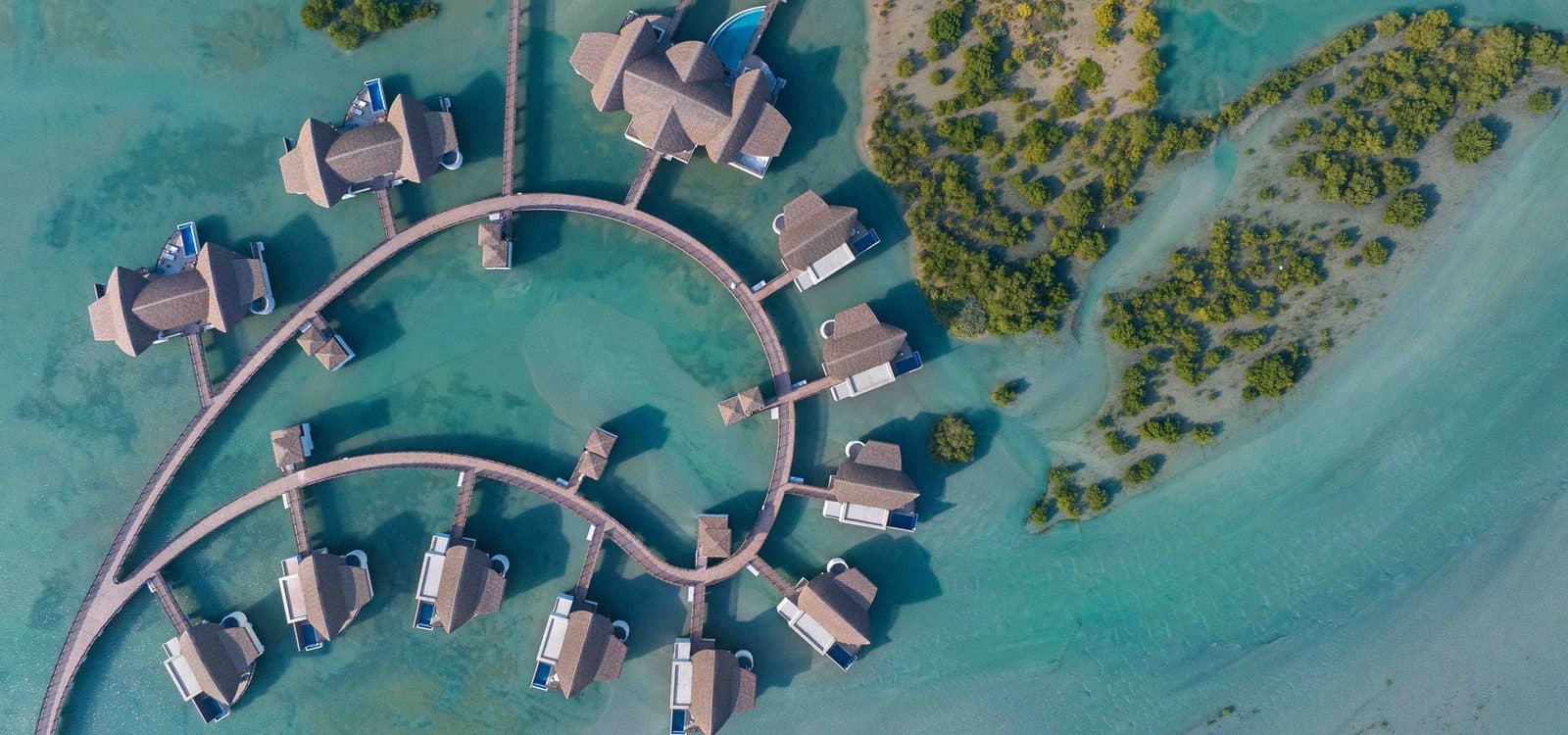 Вид сверху на виллы над водой Anantara Mina Ras Al Khaimah: дуга деревянных мостков, дома с бассейнами и мангровые заросли в бирюзовой лагуне