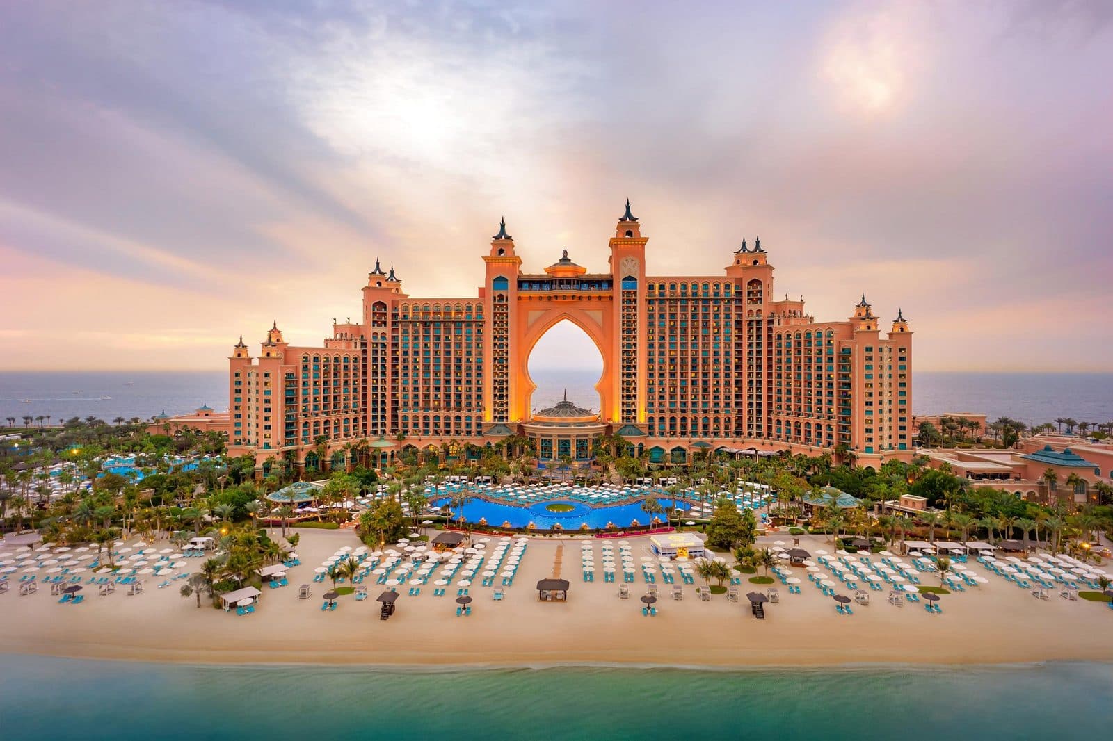 Atlantis, The Palm на закате: розовые башни с огромной аркой посередине, бассейны в пальмах и пляж с рядами лежаков у моря