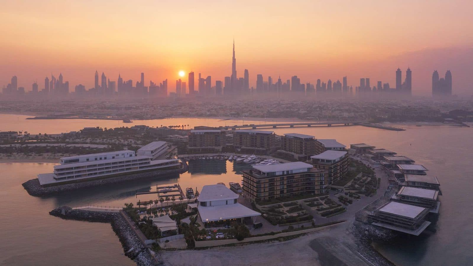 Aerial view of Bvlgari resort at sunset: buildings around a yacht marina, a bridge to the mainland and Dubai skyscrapers with the Burj Khalifa