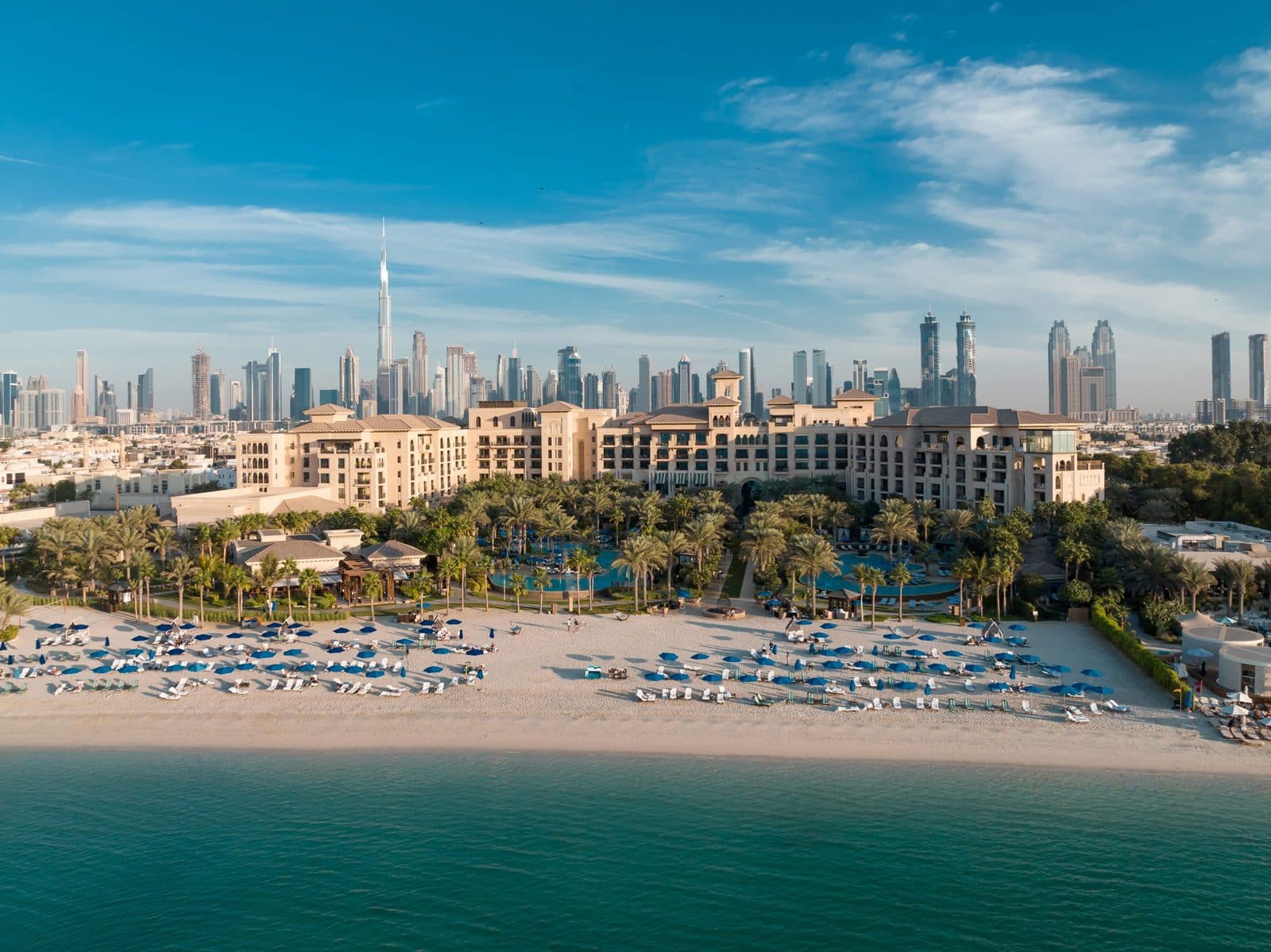 Aerial view of Four Seasons Resort Dubai at Jumeirah Beach: blue beach umbrellas, a garden with pools and sand-coloured buildings, with skyscrapers and the Burj Khalifa beyond