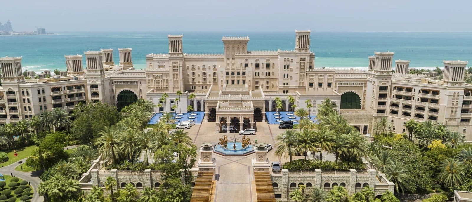 Вид сверху на Jumeirah Al Qasr: дворец с башнями-ветроловками, парадный двор с фонтанами и пальмами, за ним залив
