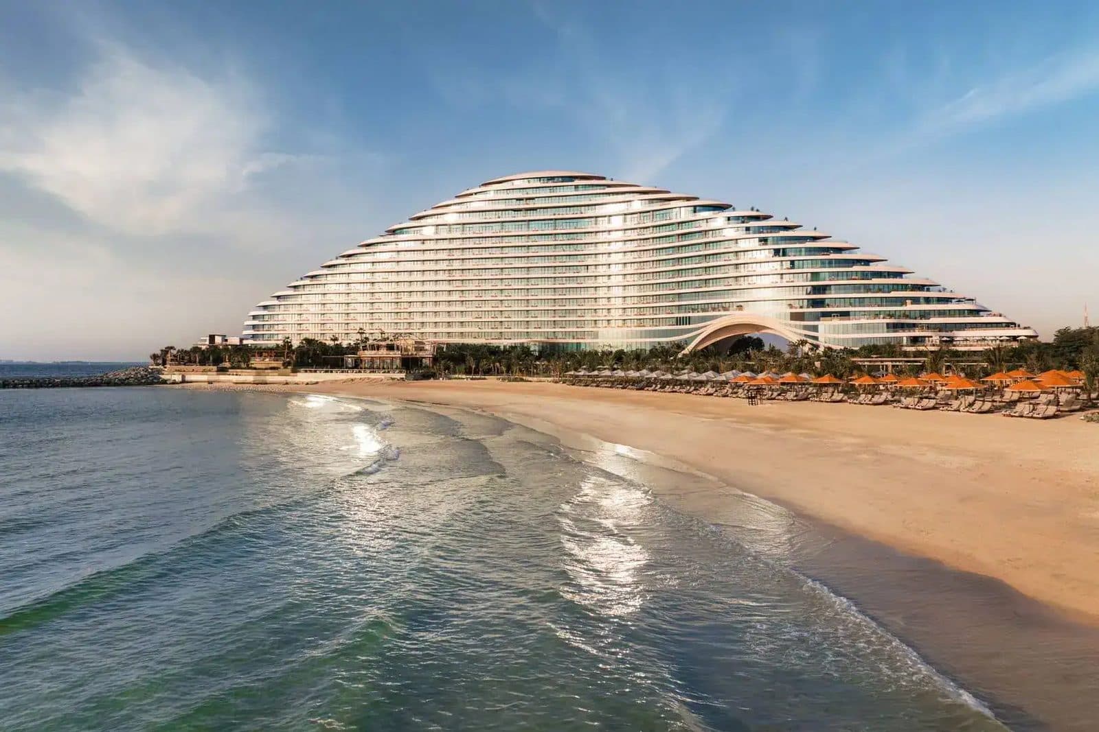 Jumeirah Marsa Al Arab с моря: волнообразное белое здание за широким песчаным пляжем с оранжевыми зонтами