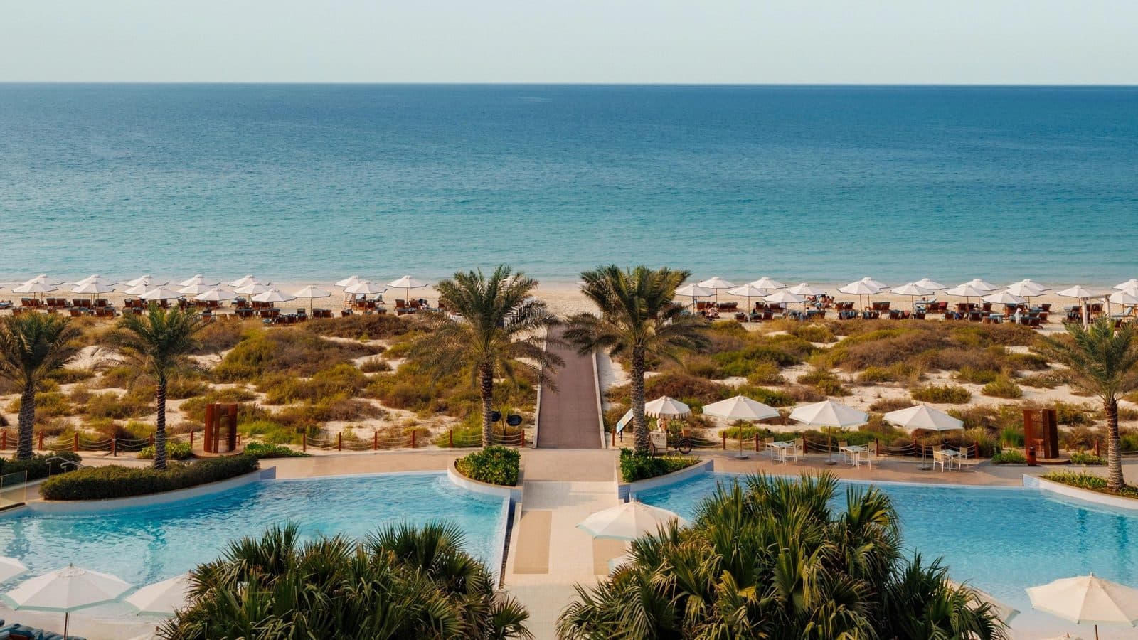 Вид с террасы Jumeirah Saadiyat Island: два бассейна среди пальм, деревянная тропа через дюны к пляжу с белыми зонтиками и Персидский залив