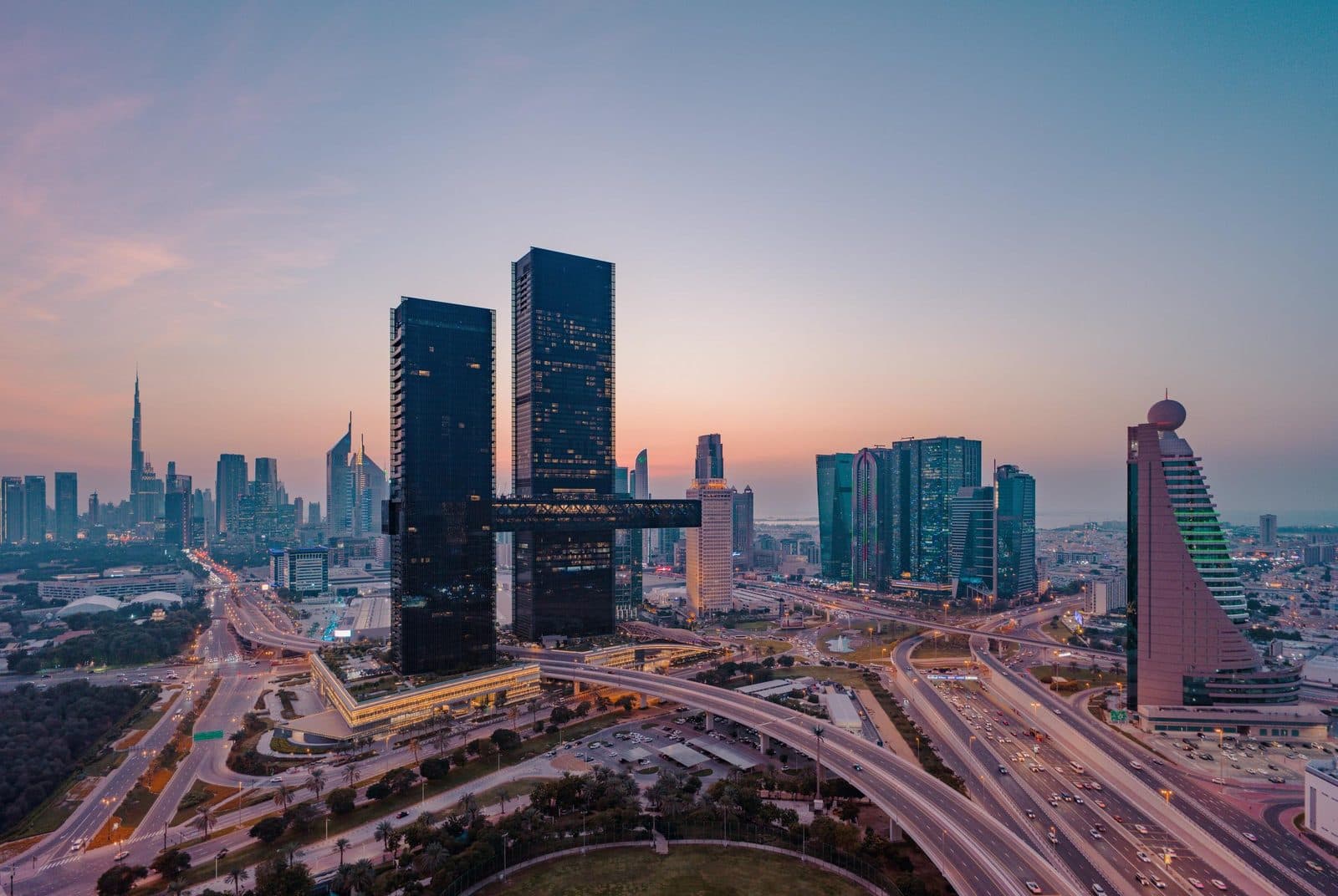Two dark One Za’abeel towers linked high above by The Link at dusk; the Burj Khalifa in the distance on the left, multi-level interchanges below