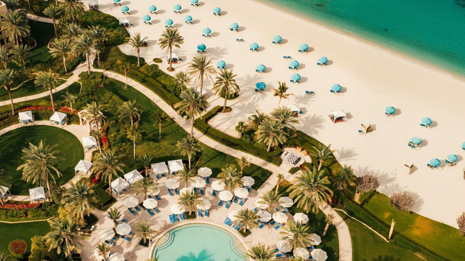 Вид сверху на One&Only Royal Mirage: пляж с бирюзовыми зонтиками, пальмовый сад с извилистыми дорожками и круглый бассейн