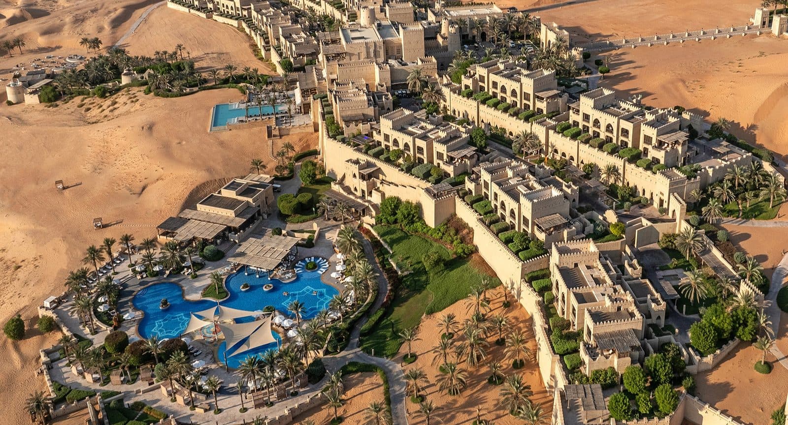 Вид сверху на Qasr Al Sarab: песочные корпуса с башнями и зубчатыми стенами, сады с пальмами и бассейн с шатрами среди дюн