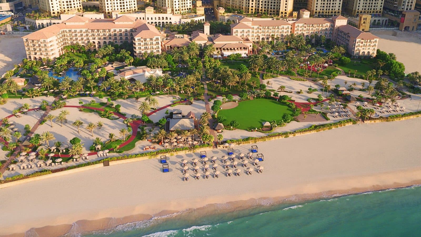 The Ritz-Carlton, Dubai сверху: терракотовые крыши корпусов среди пальм и газонов, широкий пляж с рядами зонтиков и бирюзовое море
