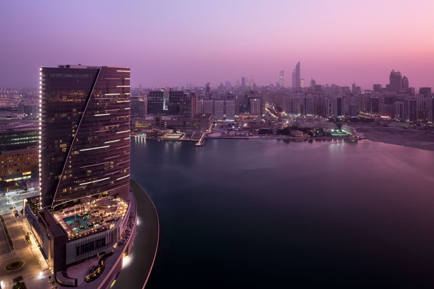 Башня Rosewood Abu Dhabi в сумерках на краю острова Аль-Марья, на другом берегу залива огни небоскрёбов Абу-Даби