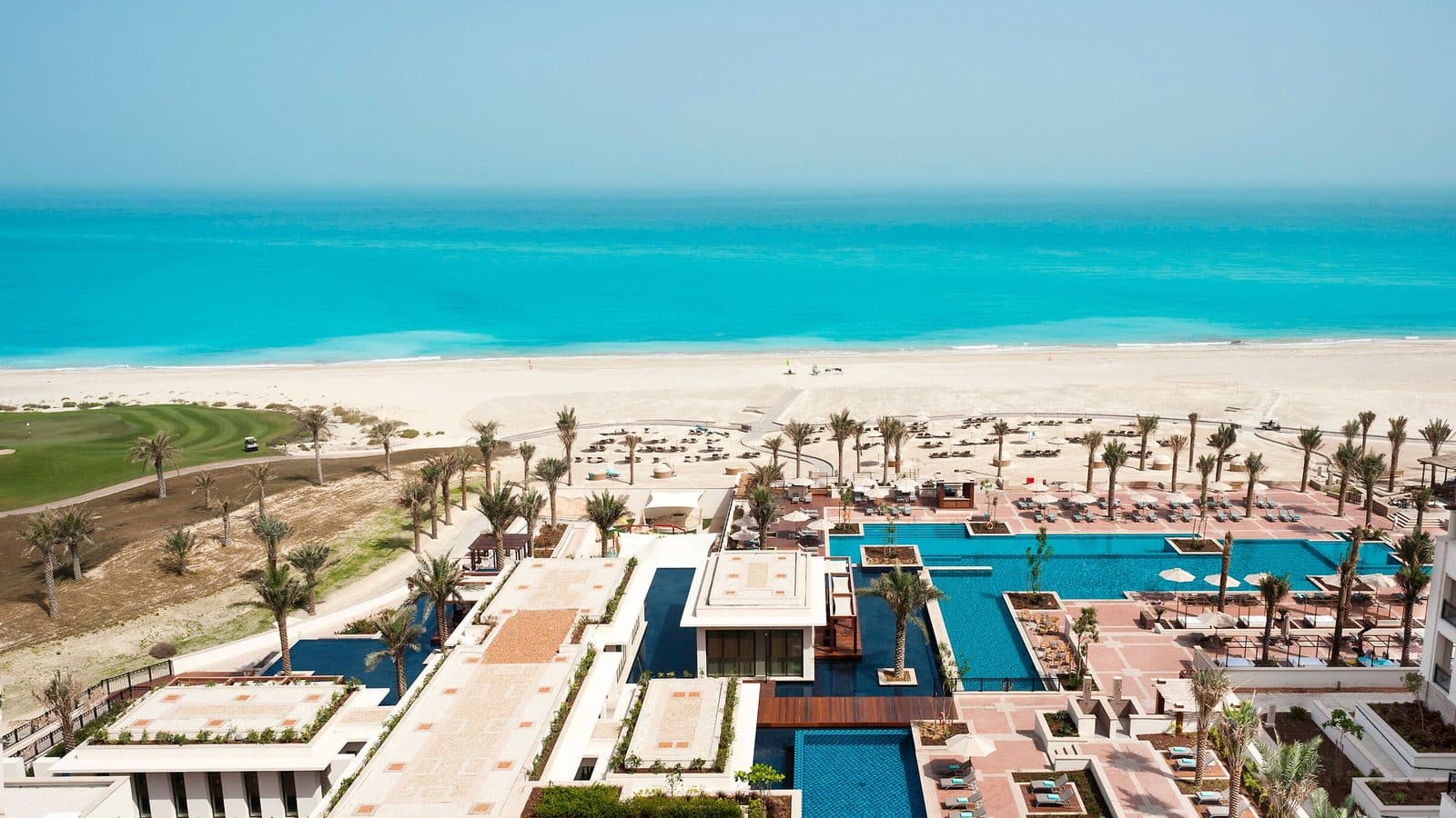 Aerial view of St. Regis Saadiyat Island: pools among palms, a broad white beach and turquoise sea, with a golf course on the left