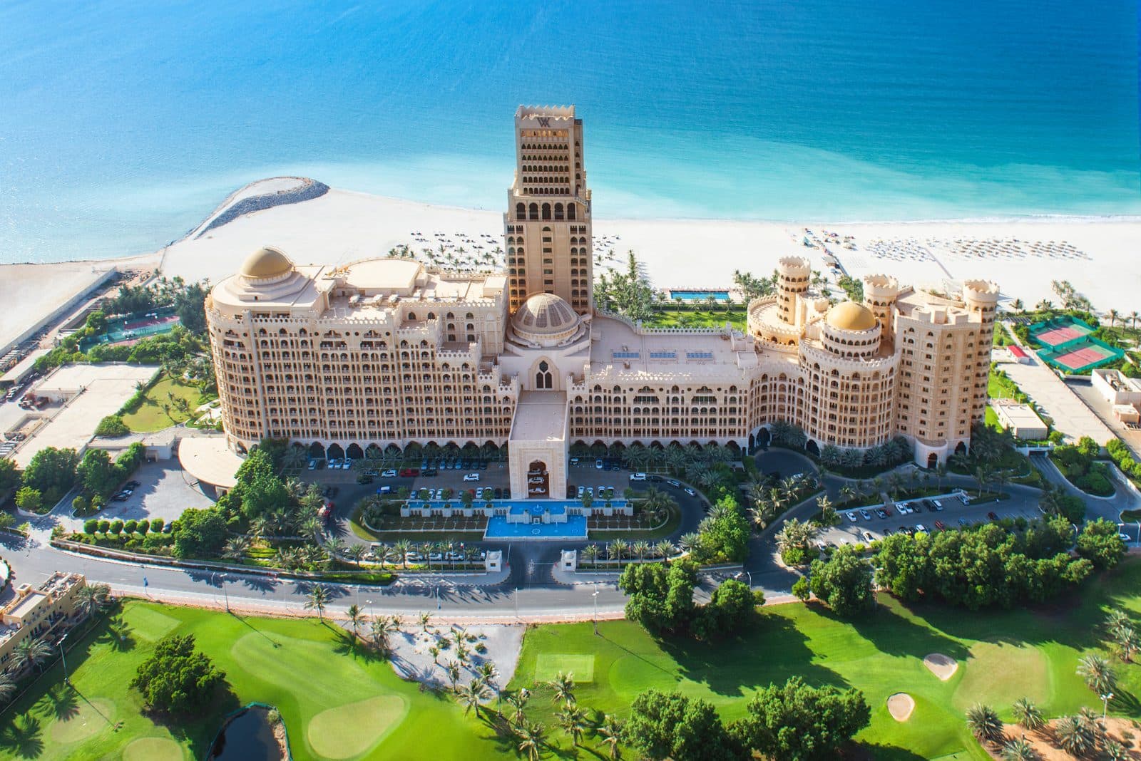 Вид сверху на Waldorf Astoria Ras Al Khaimah: здание с куполами и башней, за ним белый пляж и залив, на переднем плане поле для гольфа
