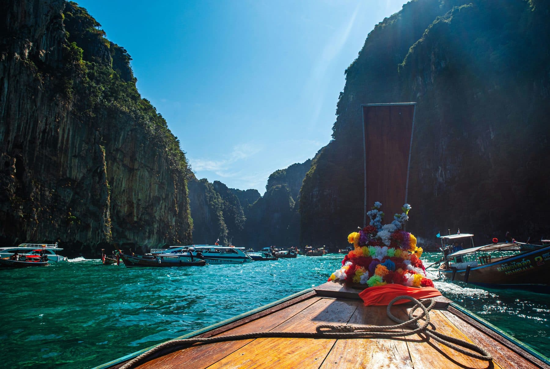 A longtail boat's bow with flower garlands enters a turquoise bay between sheer cliffs near the Phi Phi islands, with other boats ahead