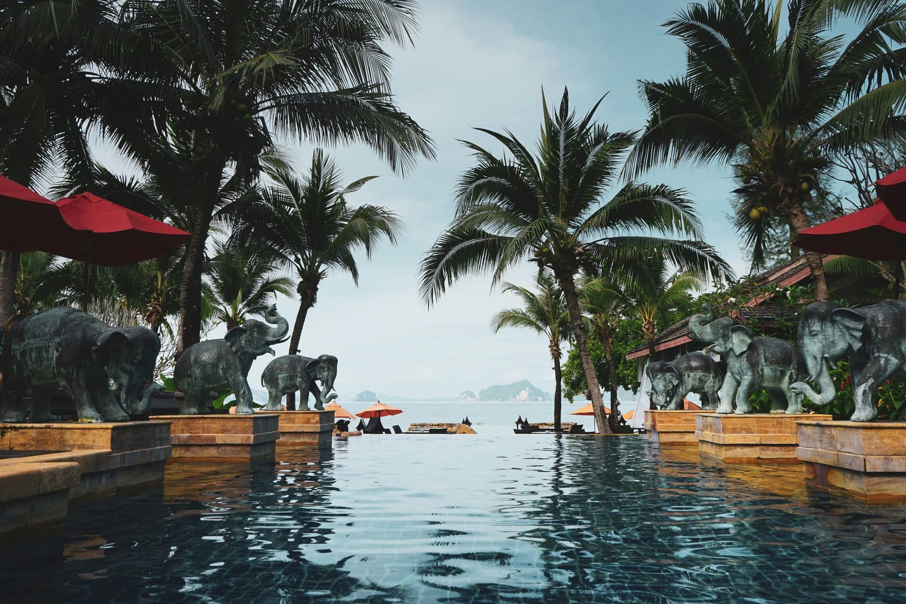 An overflow pool between palms and bronze elephant statues, with red umbrellas, beach and sea beyond and islands on the horizon; Krabi