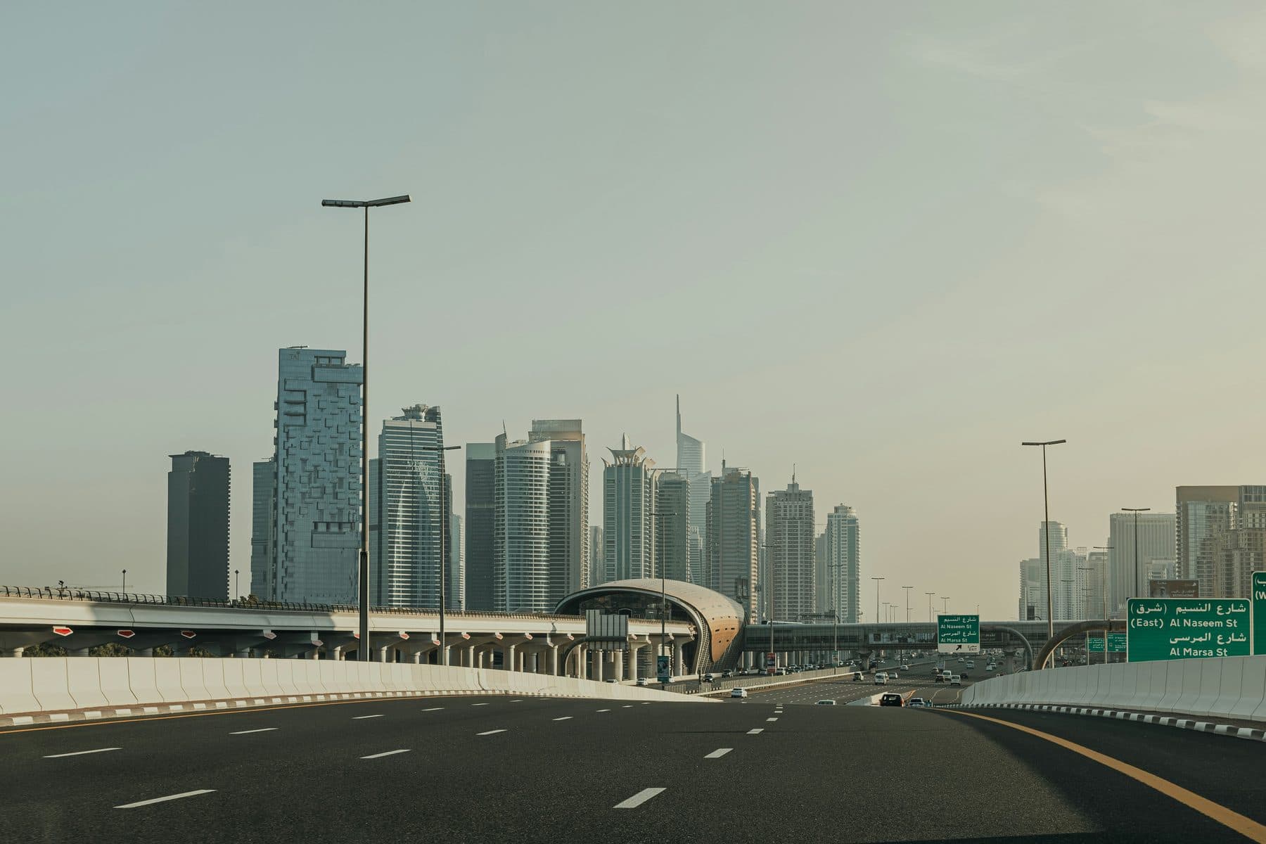 A wide, empty highway in Dubai leads towards skyscrapers; an elevated metro line and station cross the road, with green signs on the right